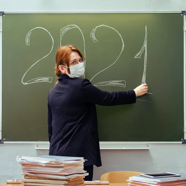 A school teacher in a medical mask writes the number 2021 in chalk on a blackboard. Learning problems during a coronavirus epidemic.