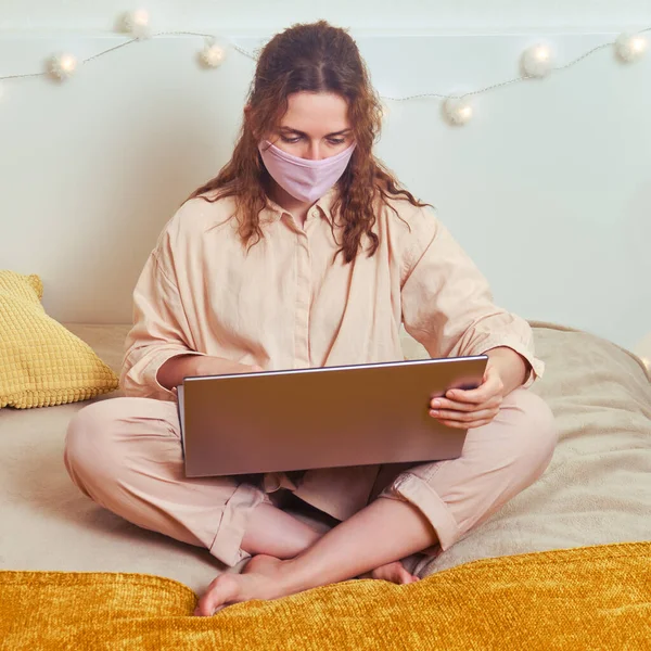 Woman in face mask works online on the yellow bed. Young girl in a medical mask with a computer at a remote work