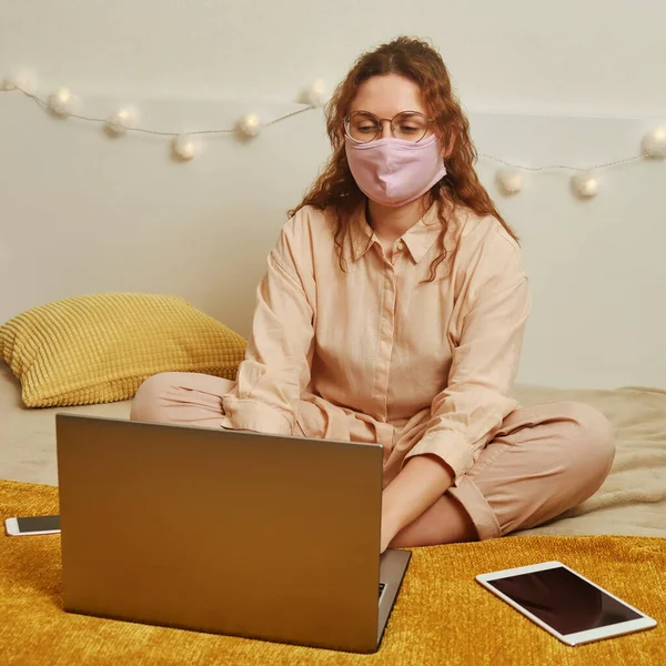 A woman in a face mask works online on a home bed. A young girl in a medical mask with a laptop for remote work