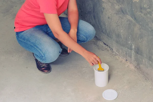 A woman squats and dips a brush into a can of paint. Painting the walls yourself in the repair of a new built house.