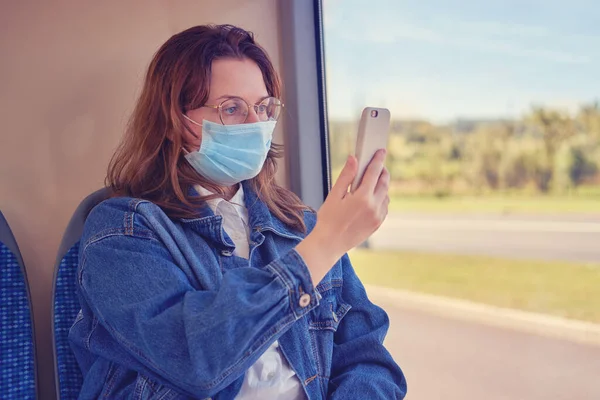 A woman in a medical mask looks at the phone sitting on a chair on the bus. A tourist takes photos on her smartphone while traveling