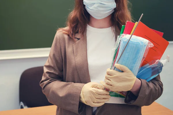 A teacher holds medical masks and a sanitizer in a school classroom