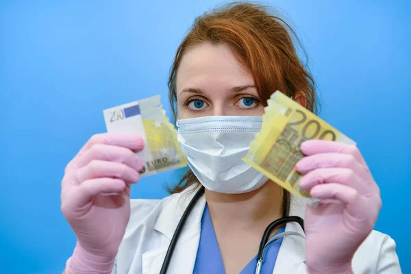 Medic holds a euro bill torn in half. A girl nurse with money for work on a blue background, the concept of paying doctors in a crisis due to the coronavirus epidemic.