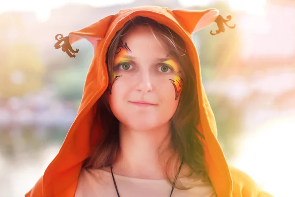 Woman in the Fox hood. Portrait of a girl with paints on her face. Squirrel ears with tassels on clothes. A woman in orange animal costume.