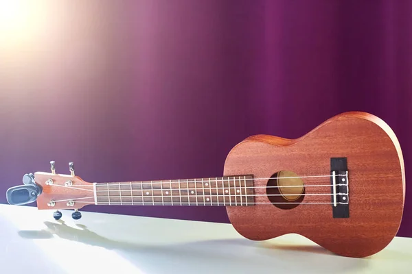 Ukulele is on a white table. Small guitar on purple background. Wooden ukulele in the sunlight.
