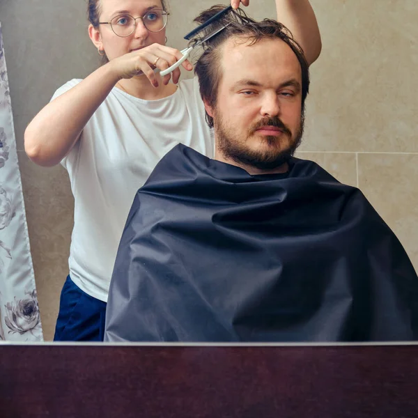 Wife cuts a man\'s hair with scissors in front of the mirror, copy space. The concept of unkempt appearance and problems in isolation from the coronavirus pandemic