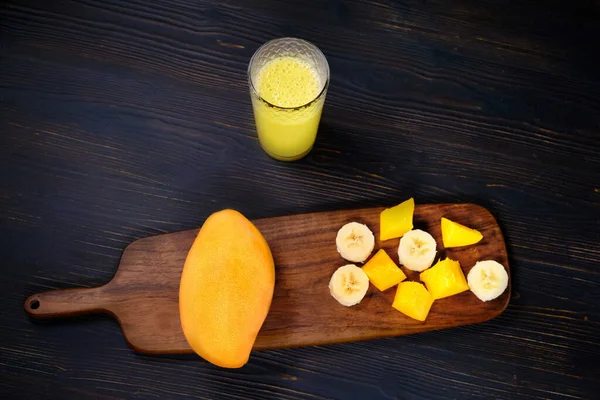 Yellow glass with fresh mango juice, copy space. Pieces of tropical fruit and a glass of smoothie. Sliced mango and banana on a dark wood background