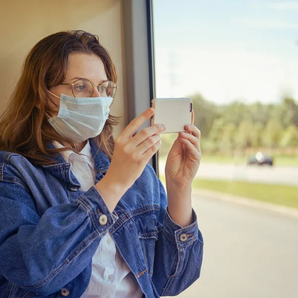 A woman in a medical mask shoots a video on the phone in a transport. Trips on public bus during an epidemic due to the flu virus