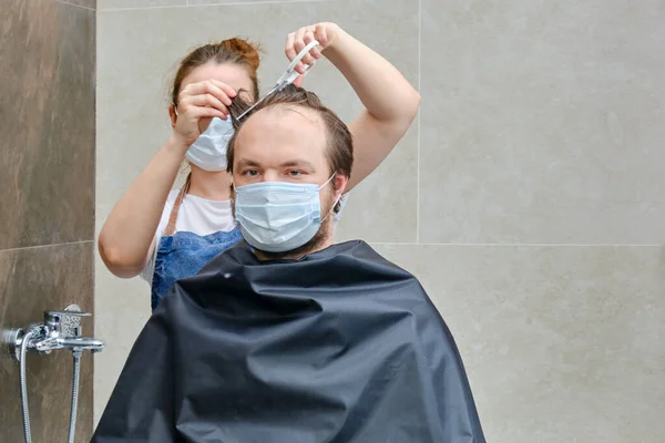A woman in medical mask cuts a man's hair with scissors, copy space