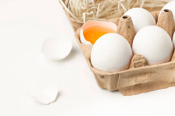 macro shot of white eggs in carton on the white background. One egg is cracked with the yolk and white exposed. White shell is laying around.