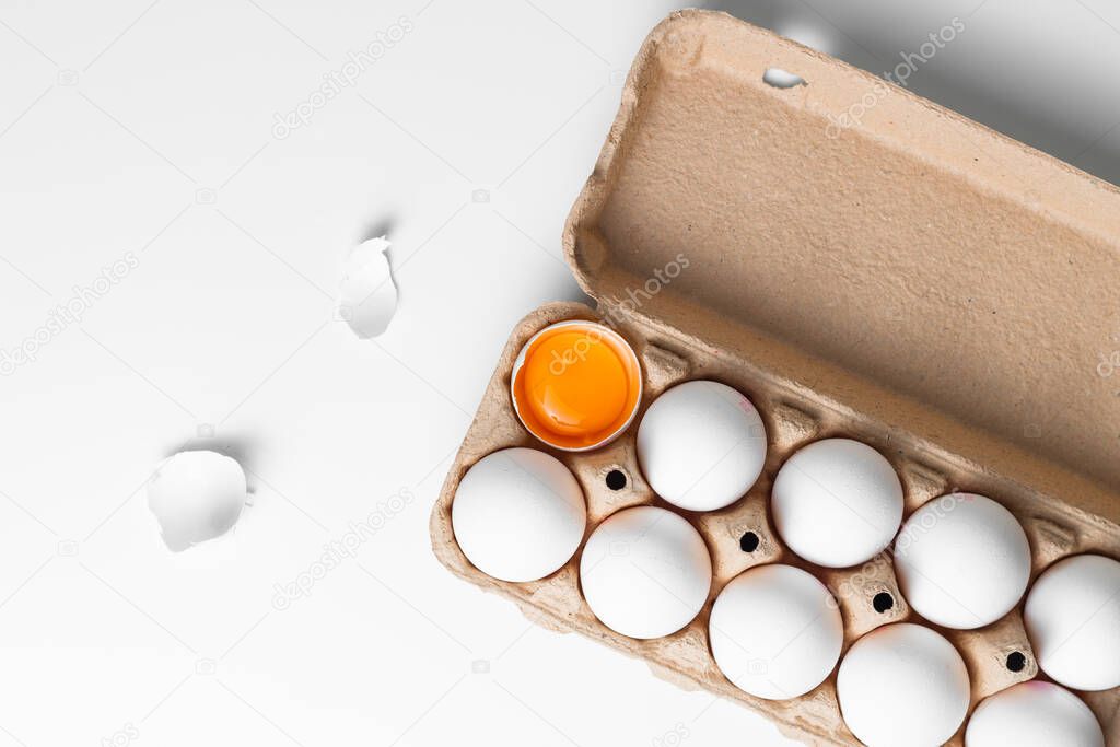 Overhead view of white eggs in carton on the white background. One egg is cracked with the yolk and white exposed. White shell is laying around.