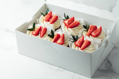 Çilekle süslenmiş beyaz kremalı çikolatalı kekler. Beyaz mermer arka plandaki beyaz hediye kutusuna istiflenmiş çörekler.