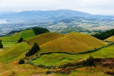 Sao Miguel Adası 'ndaki yeşil tepelerin manzarası, Azores, uzak bir kasaba ve bulutlu bir gökyüzünün altındaki dağlar..