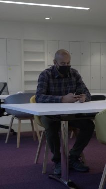 Yetişkin erkek maske takıyor. Akıllı telefon kullanıyor. Geniş masada oturuyor. Dikey Video, Kilitli