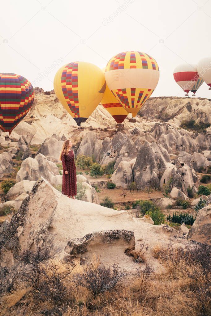 Beautiful woman enjoying amazing view and walking in Cappadocia landscape with hot air balloon on background at sunrise. Travel in Turkey
