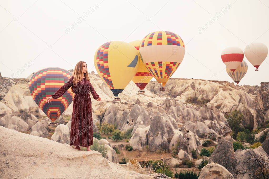 Beautiful woman enjoying amazing view and walking in Cappadocia landscape with hot air balloon on background at sunrise. Travel in Turkey