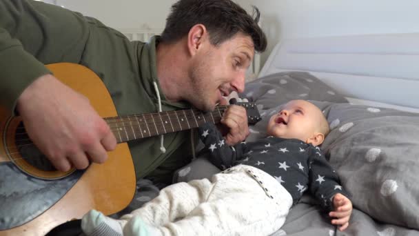 Seorang ayah yang menarik bermain gitar untuk bayi laki-lakinya yang menggemaskan di tempat tidur — Stok Video