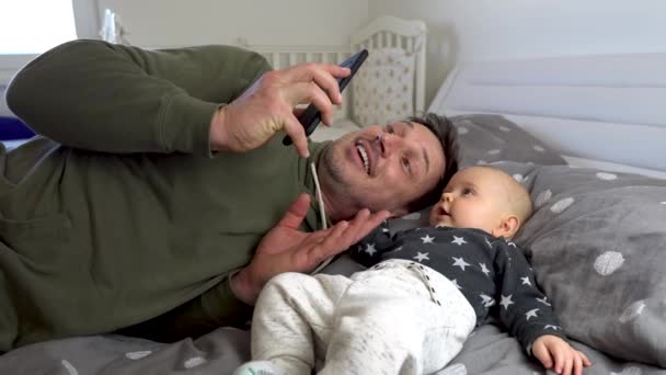 Lykkelig far en søt gutt som ser ut som en mamma på en mobiltelefon i en seng. 4K – stockvideo
