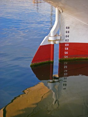 Metal tekne dümeni kısmen denize batmış ve suya yansıyor. Renkli beyaz ve kırmızı.