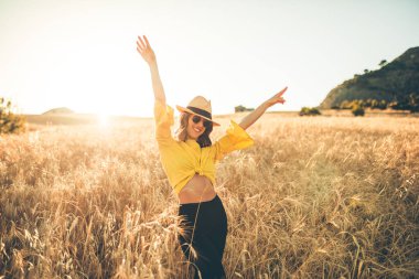 Sarı şapkalı genç kız gün batımında buğday tarlasının ortasında mutlu duruyor..