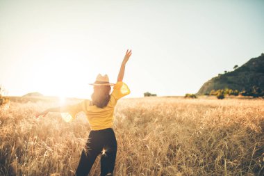 Sarı şapkalı genç kız gün batımında buğday tarlasının ortasında mutlu duruyor..
