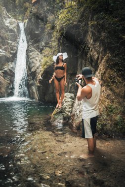 Fotoğrafçı, şelalenin karşısında beyaz şapkalı genç bir kadının fotoğraflarını çekiyor.