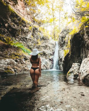 Şelalenin karşısında poz veren şapkalı genç kadın, kameraya doğru suyun içinde duruyor.
