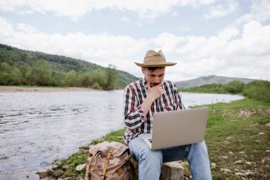 Çevrimiçi öğrenim gören bir adam dışarıda, dağlarda dizüstü bilgisayarla öğreniyor.