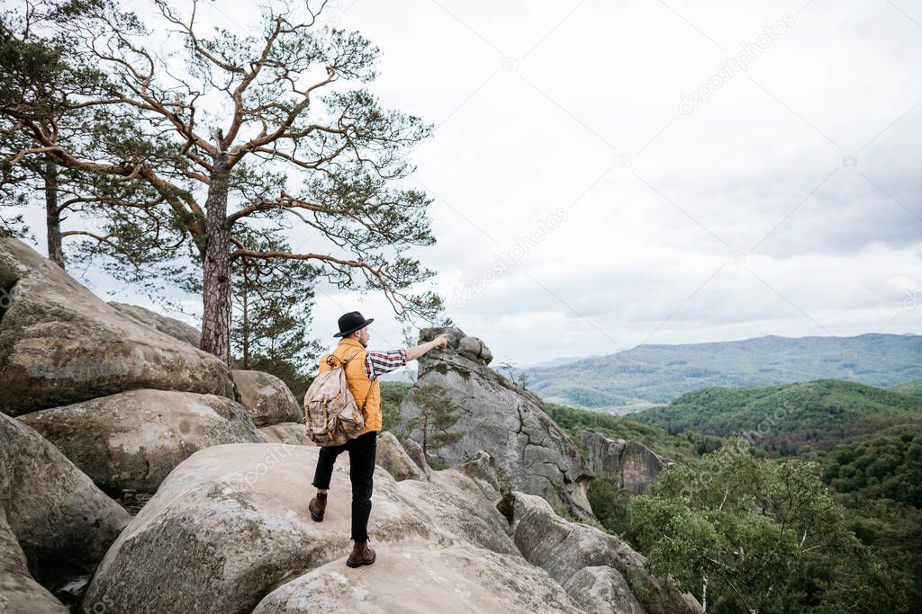 A young hipster hiker stands on top of a cliff among a mountain forest