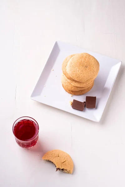 Homemade stack of shortbread chocolate cookies with one bitten cookie and two pieces of chocolate on white plate with stewed fruit red compote drink in glass on wooden table