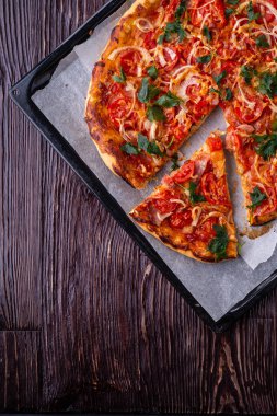 Taze pişmiş sıcak pizza ve dilim peynir jambon et domates soğan maydanoz ev yapımı parşömen kağıt üzerinde fırın levha tepsi tarifi koyu ahşap masa üstte görünümü