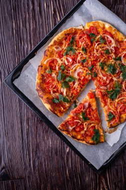 Taze pişmiş sıcak pizza ve dilim peynir jambon et domates soğan maydanoz ev yapımı parşömen kağıt üzerinde fırın levha tepsi tarifi koyu ahşap masa üstte görünümü