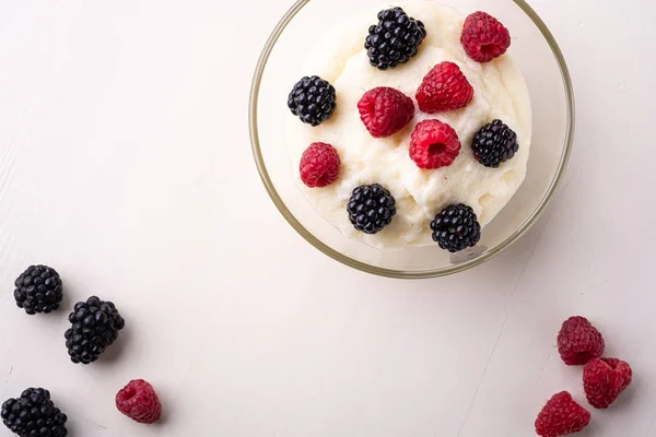 Semolina pudding porridge dessert with blackberry and raspberry in glass bowl near with scattered heap berries on white wooden background, breakfast, isolated, top view