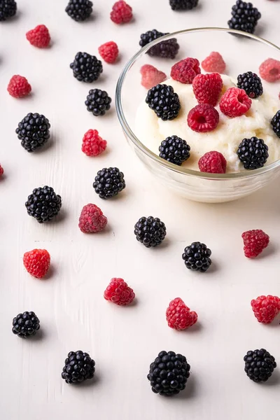 Semolina pudding porridge dessert with blackberry and raspberry in glass bowl near with scattered heap in order berries on white wooden background, breakfast, isolated, selective focus