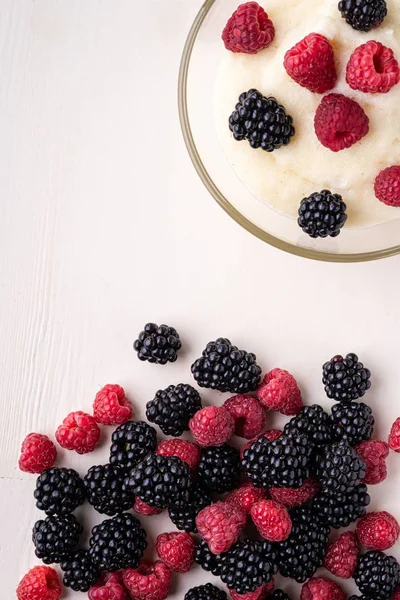 Semolina pudding porridge dessert with blackberry and raspberry in glass bowl near with scattered heap berries on white wooden background, breakfast, isolated, top view