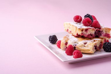 Ev yapımı kek parçası, ahududu ve böğürtlen meyveleri ile iki pasta dilimleri, pudra şekeri, pembe kağıt arka plan üzerinde beyaz kare plaka