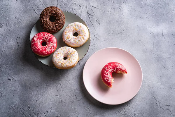 Bitten pink donut with sprinkles on pink plate near to chocolate and vanilla donuts, sweet glazed dessert food on concrete textured background, angle view