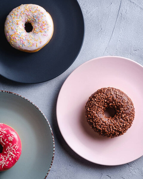 Three donuts on plates, chocolate, pink and vanilla donut with sprinkles, sweet glazed dessert food on concrete textured background, top view