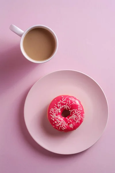 コーヒーのカップの近くのプレート上にスプリンクルとピンクのドーナツ 甘いガラスのデザートフードとピンクの最小限の背景に熱い飲み物 トップビュー — ストック写真
