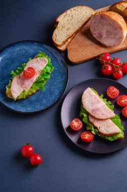 Hindili sandviç, yeşil salata ve taze vişneli domates dilimleri kesim tahtasındaki malzemelerin yanında, mavi minimal arka plan, açı açılı makro.