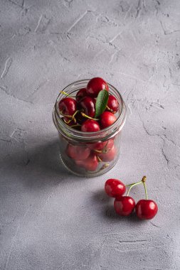 Taze olgun kiraz dalı, cam kavanozda yeşil yapraklar olan kirazların yanında, gri taş arka planda yaz vitamini meyveleri, açı açılı makro.