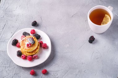 Süzme peynir krepleri ve pudra şekeri, kremalı börek, ahududu ve böğürtlenli tatlı tabakta, beton zemin üzerinde limon dilimi olan sıcak çay fincanının yanında, açıyla kopyalama alanı