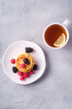 Süzme peynirli krep, kremalı pasta ahududu ve böğürtlenli tatlı tabakta sıcak çay bardağının yanında taş zeminde limon dilimleri, üst manzara.