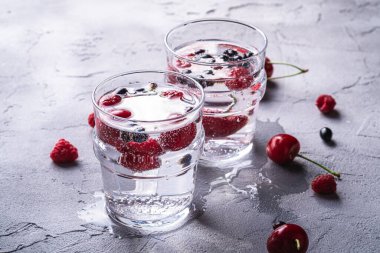 Taş zeminde iki şeffaf bardakta kiraz, ahududu ve frenk üzümlü taze, soğuk, köpüklü su içeceği yaz diyeti içeceği, açılı makro görüş.