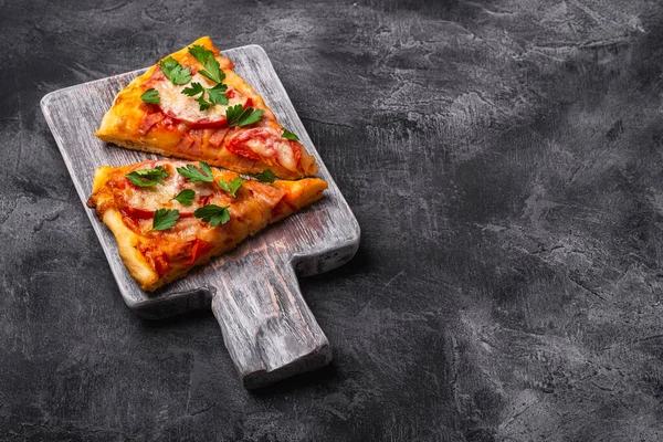 Hot pizza slices with mozzarella cheese, ham, tomato and parsley on wooden cutting board, stone concrete background, angle view copy space for text