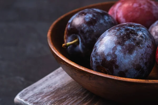 Kahverengi ahşap kasede taze erik meyveleri eski kesim tahtasının üzerinde, siyah desenli arka plan, açı açılı makro.