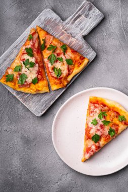 Sıcak pizza dilimleri mozzarella peyniri, jambon, domates ve maydanoz ahşap kesim tahtası ve tabak, beton arka plan, üst manzara