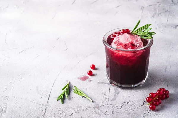 Fresh ice cold fruit cocktail in glass, refreshing summer red currant berry drink with rosemary leaf on stone concrete background, angle view copy space