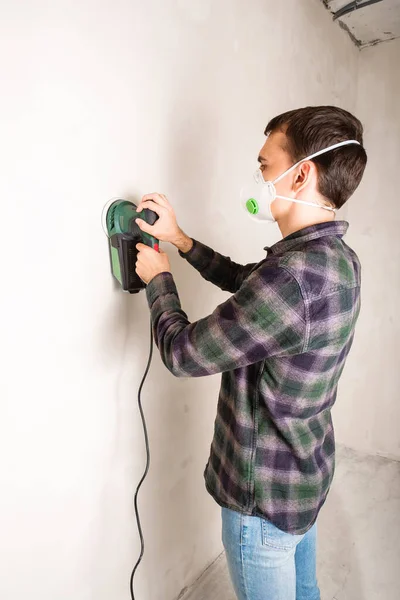 Man in protective mask working with electric sander to smooth plaster wall surface, room renovation concept