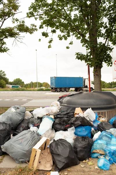 Yol kenarındaki konteynırları dolduran siyah ve renkli çöp torbaları. Karton ve plastik ambalajlar atık malzemeler arasında görünür.